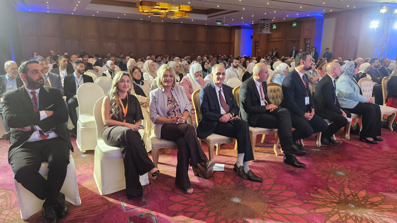 Group of professionals in business attire seated in rows at a conference hall, wearing lanyards and facing the stage.