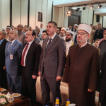 Group of men standing in a formal conference hall, with a flower-adorned table and snacks in the foreground.