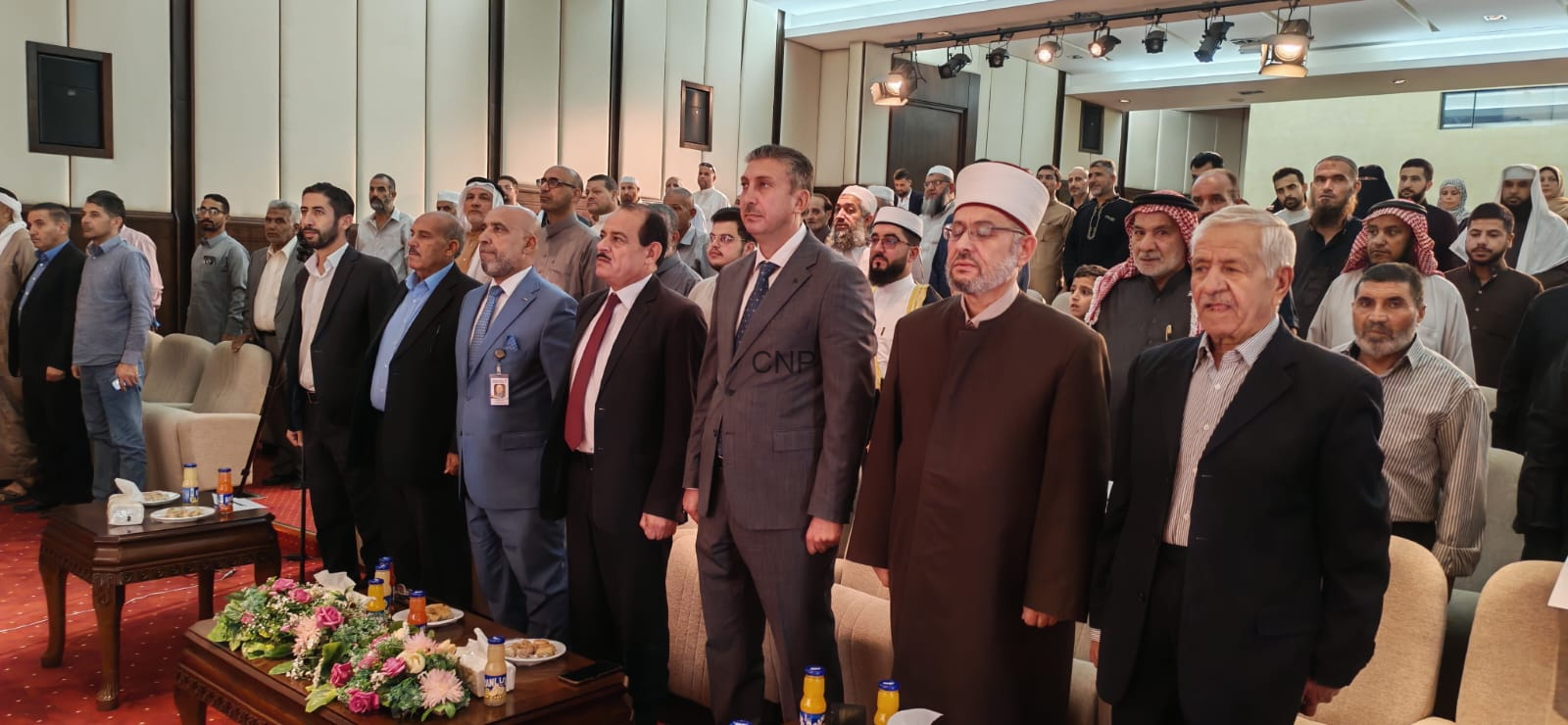 Group of men standing in a formal conference hall, with a flower-adorned table and snacks in the foreground.
