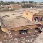 Aerial view of a damaged beige building with a collapsed roof and rubble on the terrace, with palm trees and surrounding ruined structures in the area.