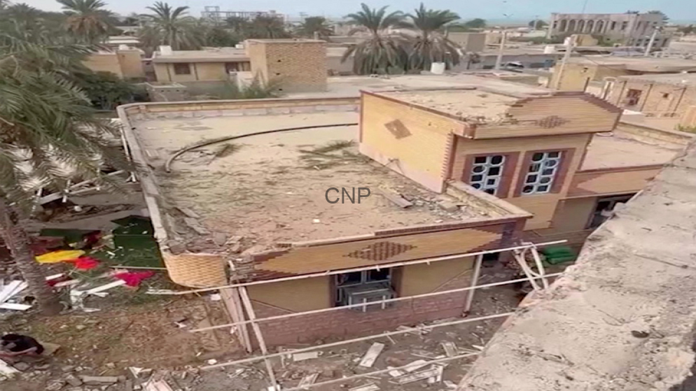 Aerial view of a damaged beige building with a collapsed roof and rubble on the terrace, with palm trees and surrounding ruined structures in the area.