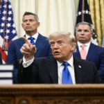 Man in a dark suit and blue tie points with his index finger while seated at a desk, flanked by four suited men and U.S. flags in the background.