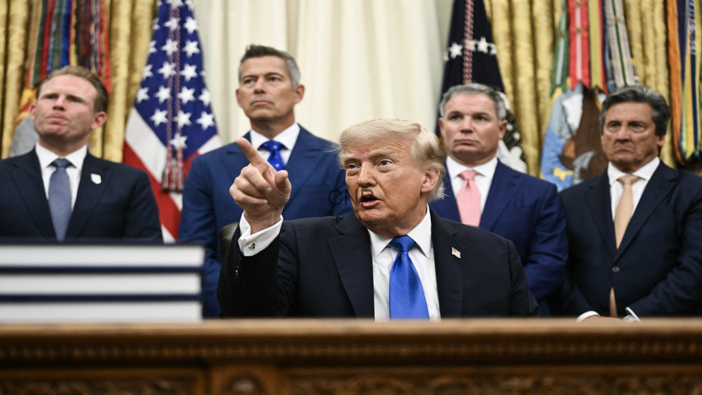 Man in a dark suit and blue tie points with his index finger while seated at a desk, flanked by four suited men and U.S. flags in the background.