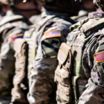 Line of soldiers in camouflage gear with American flag patches, standing at attention.