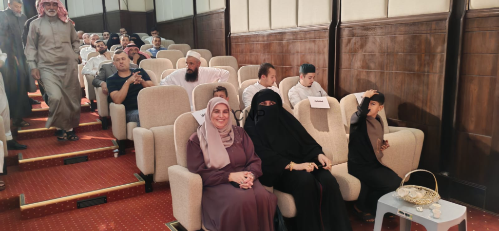 image - Center News Network Audience seated in a red-carpeted auditorium; front row features a smiling woman in a purple dress and hijab beside a woman in a black niqab, with a boy to their right.