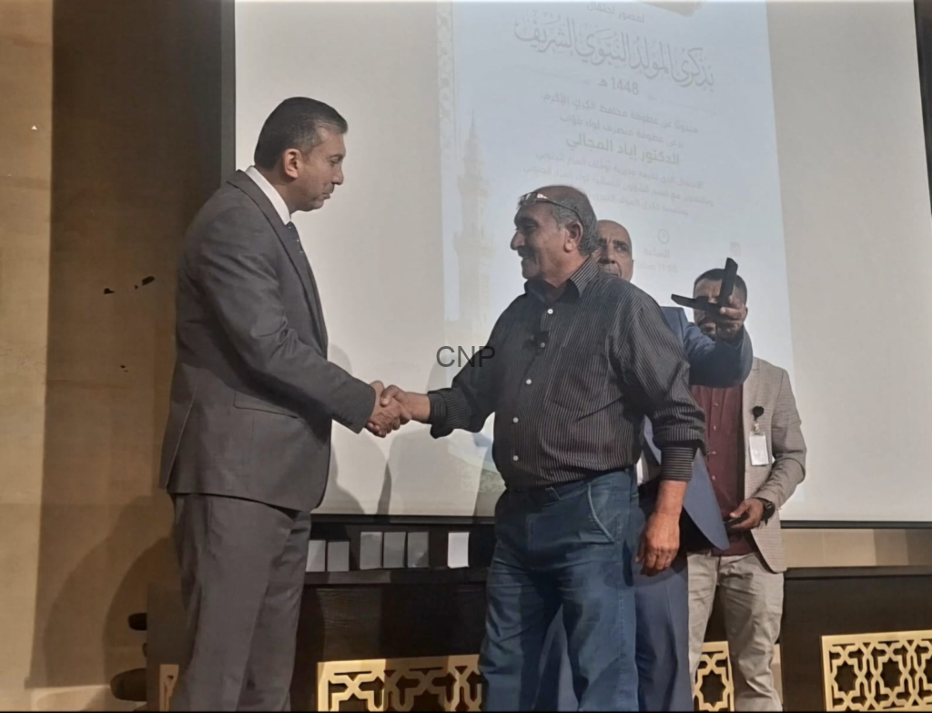 Two men shake hands on a stage during a ceremony, with a projection screen behind them.
