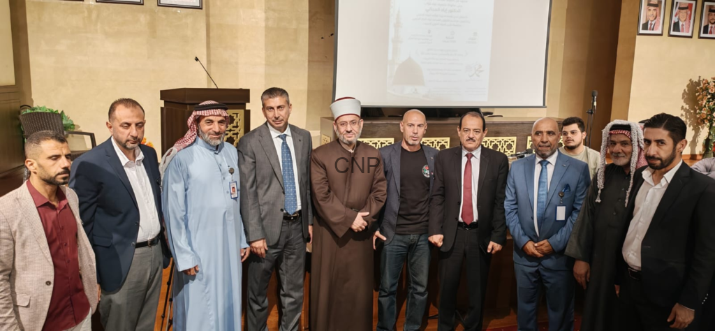 Group of men in formal attire posing for a photo in a conference hall, central figure in brown traditional robe and white turban.
