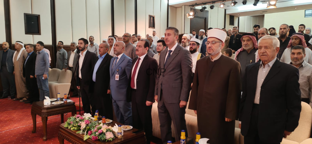 image - Center News Network Group of men in suits and traditional robes stand in a conference hall during a formal ceremony, with flowers and beverages on the tables in front.
